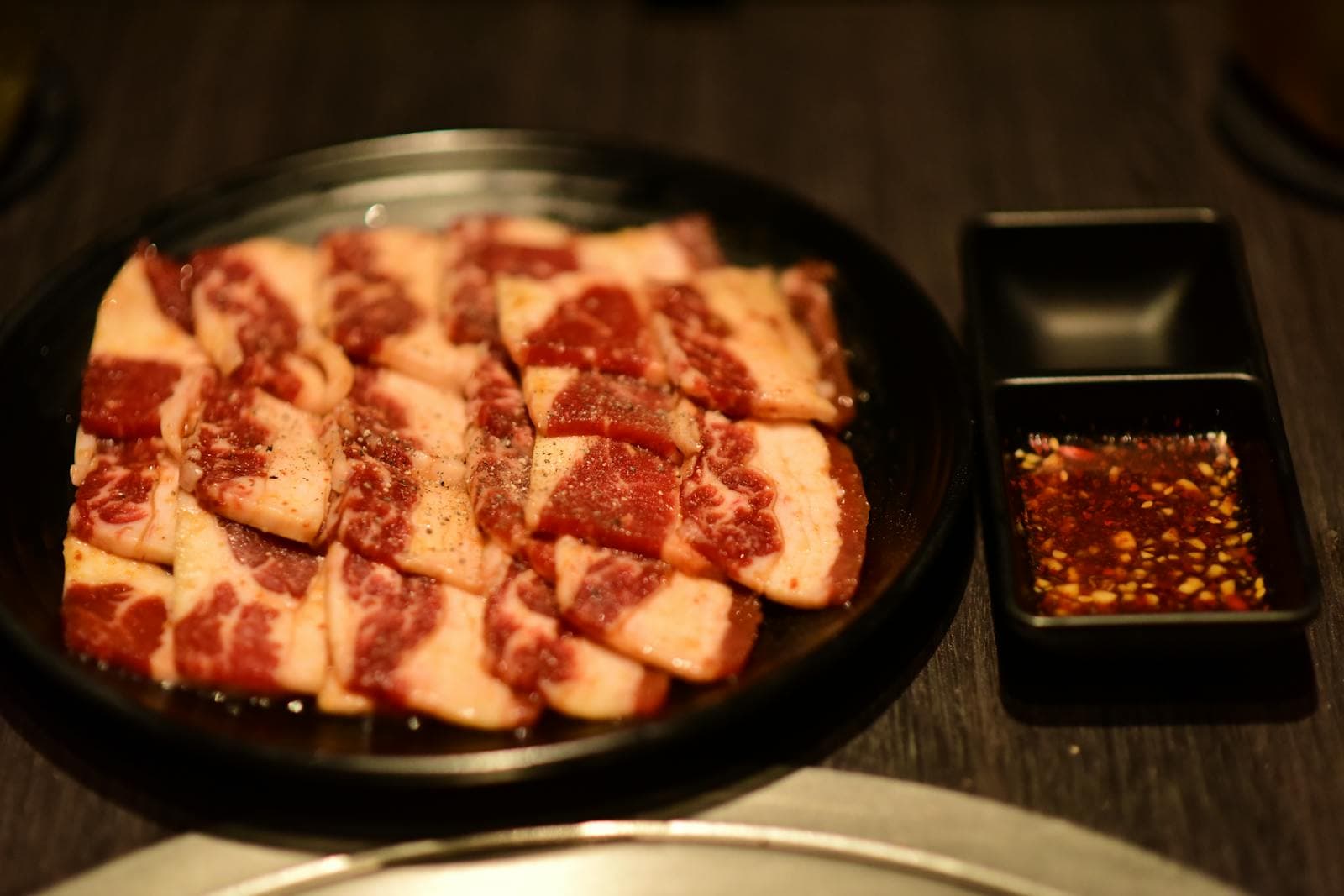 Square cuts of marbled galbisal hanwoo beef on a black plate at a Korean BBQ restaurant, with chili-flake dipping sauce on the side