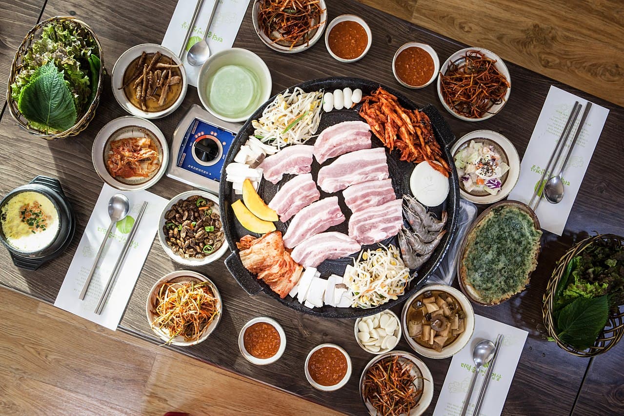 Overhead shot of a Korean BBQ table with samgyeopsal pork belly slices on a circular grill, surrounded by banchan dishes, ssam vegetables, and dipping sauces