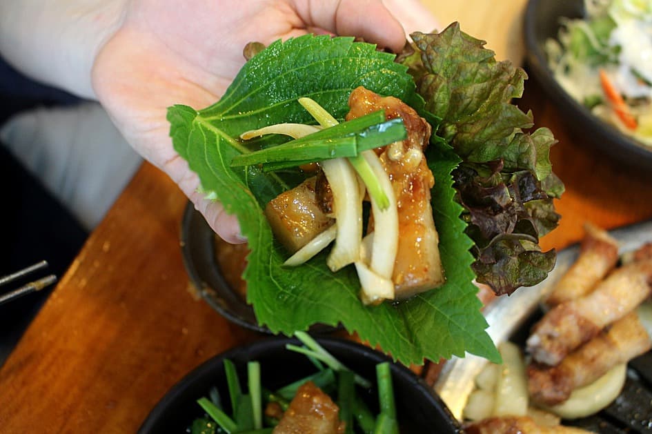 A Korean ssam wrap built in an open palm: red leaf lettuce holding a perilla leaf, a piece of grilled pork belly, slices of garlic, scallions, and a dab of ssamjang