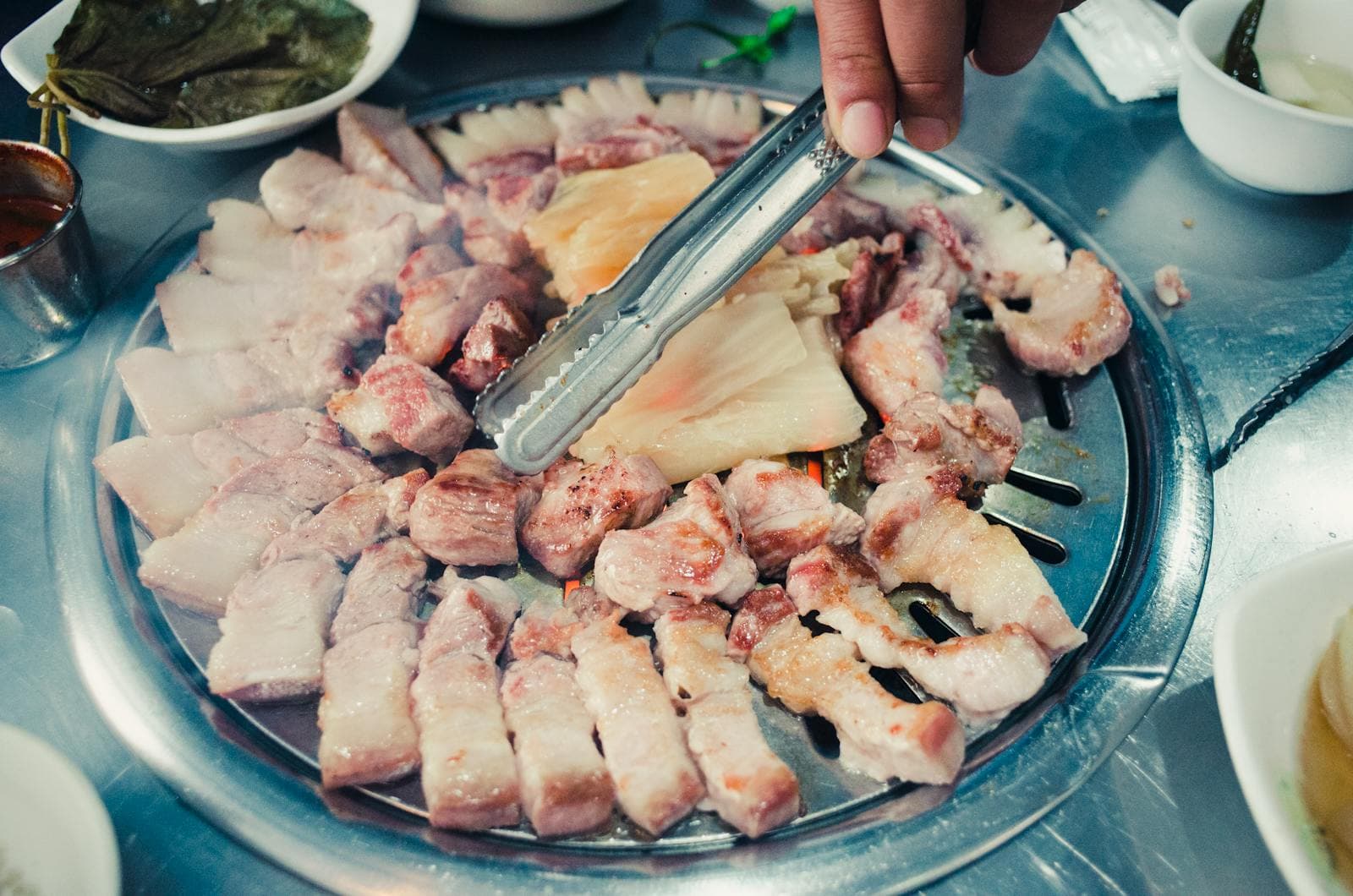 A hand using stainless-steel tongs to handle slices of samgyeopsal pork belly on a circular Korean BBQ grill